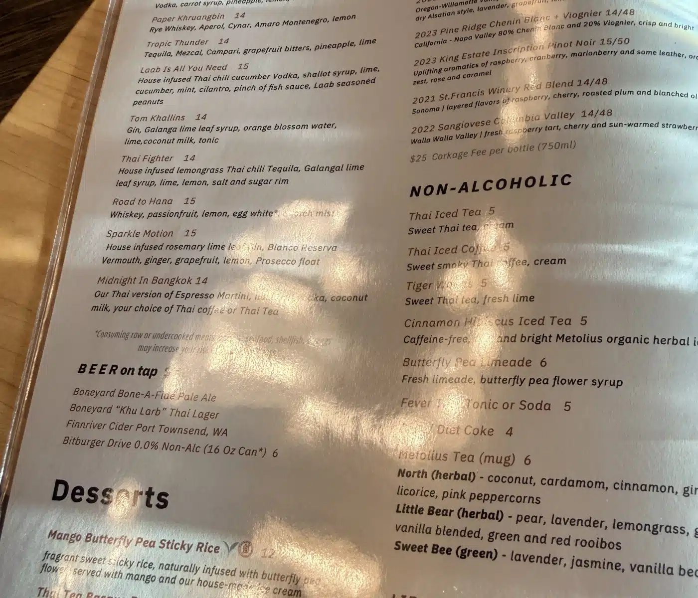 a Sen Thai Noodles Menu Printed on Textured Off white Cardstock Lists Sections for Cocktails Wines Non alcoholic Drinks and Desserts the Text is Printed in Dark Grey with Elegant Headers and the Menu is Angled Slightly on a Dark Wooden Surface
