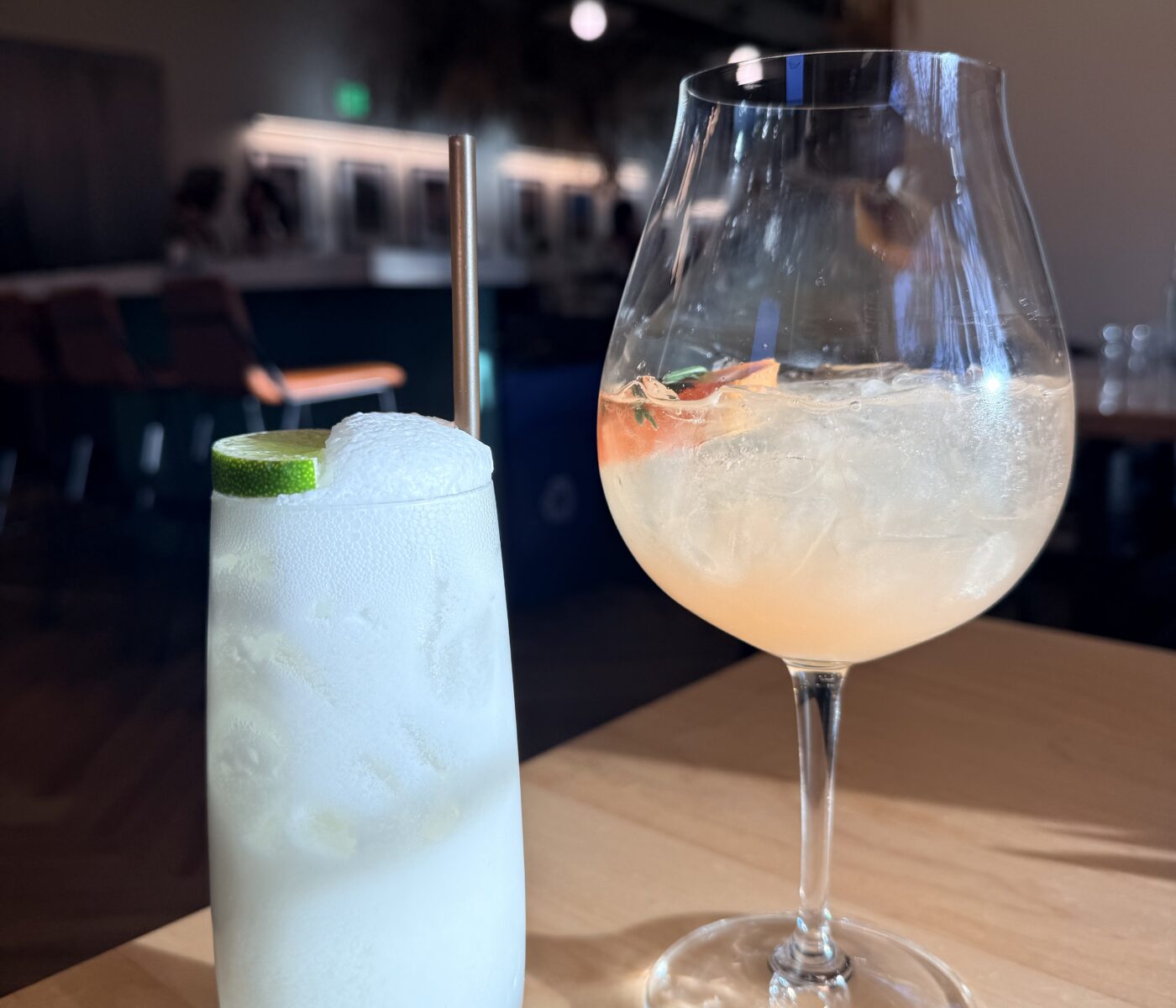 Two Cocktails at Sen Thai Noodles Sit on a Light Wood Table in a Dim Restaurant on the Left is a Frothy White Drink with a Lime Slice and Metal Straw and on the Right is a Pale Pink Spritz in a Stemmed Wine Glass Filled with Ice and a Citrus Garnish