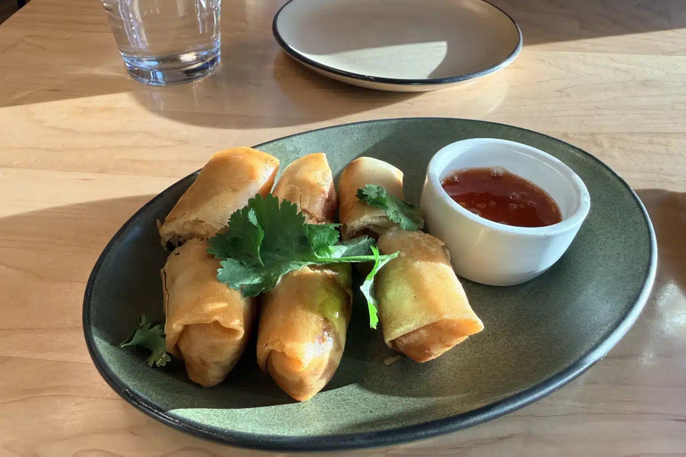 Four Golden Brown Sen Thai Noodles Spring Rolls Are Arranged on a Dark Green Oval Plate with a Small Side of Reddish Dipping Sauce and a Sprig of Cilantro the Plate Sits on a Light Wooden Table Next to an Empty White Plate and a Glass of Water
