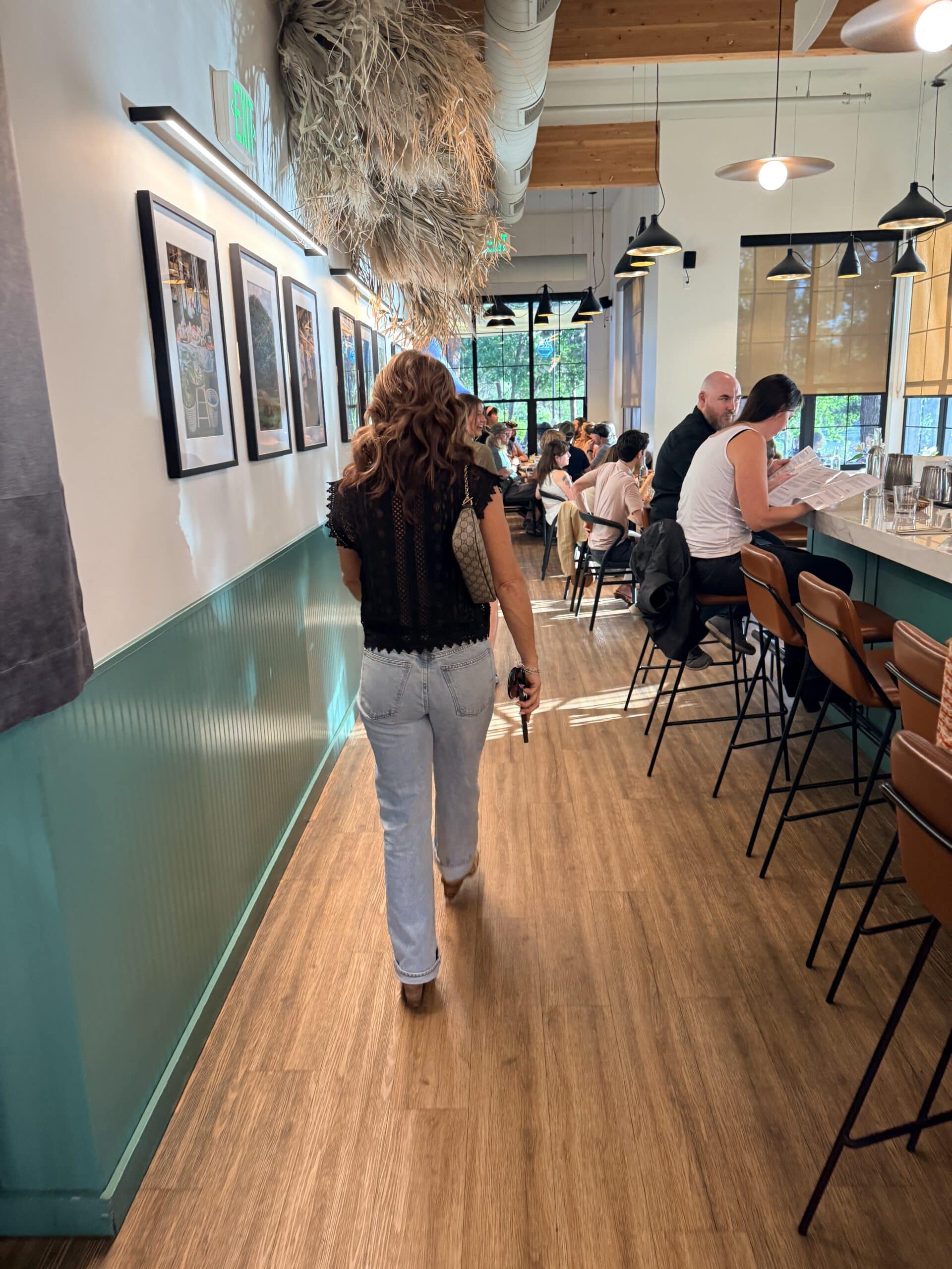 a Woman with Wavy Hair Walks Away from the Camera Through the Bright Sen Thai Noodles Cafe Wearing a Black Lace Top and Light Wash Jeans to Her Right Patrons Sit at a Long Counter on Brown Bar Stools While Framed Pictures Hang on the Teal and White Wall to Her Left