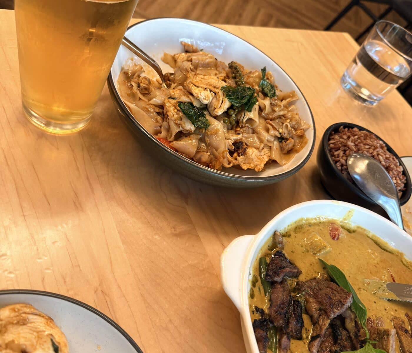 a High Angle View Shows a Meal on a Light Wood Table Including a Bowl of Sen Thai Noodles with Chicken a Dish of Green Curry with Beef a Glass of Beer and a Small Bowl of Brown Rice a Partial Plate of Fried Food Sits in the Foreground and a Glass of Water is Nearby