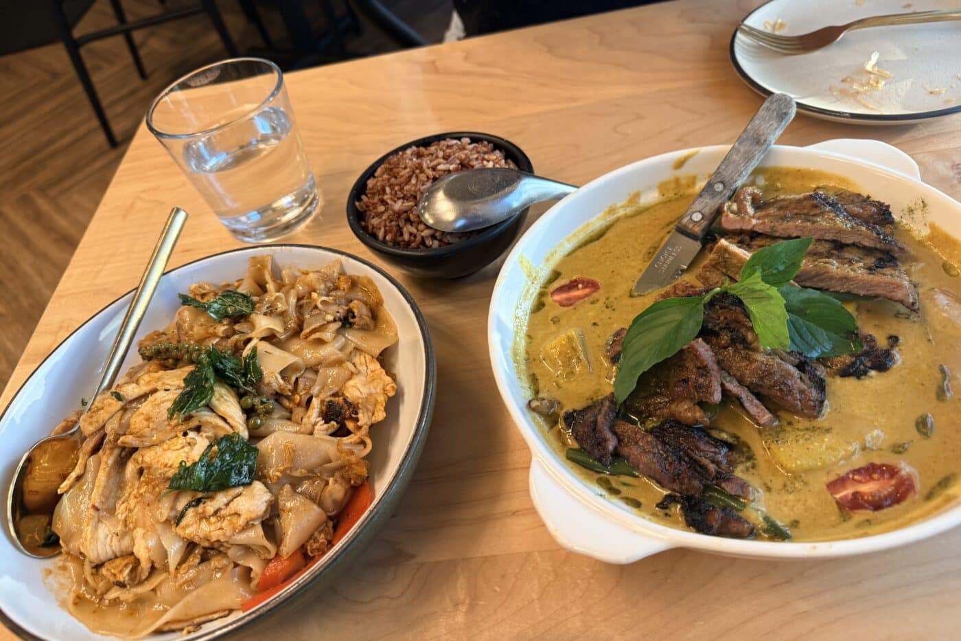 a Wooden Table is Spread with Thai Dishes Including a Bowl of Sen Thai Noodles with Chicken a Bowl of Red Rice and Sliced Grilled Meat in Green Curry the Curry is Topped with Fresh Basil and a Glass of Water Sits Nearby Next to a Used Plate and Fork