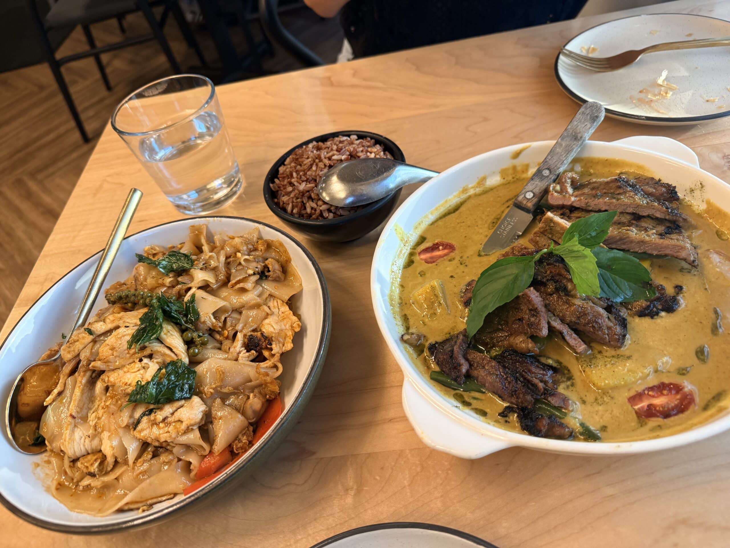 a Wooden Table is Spread with Thai Dishes Including a Bowl of Sen Thai Noodles with Chicken a Bowl of Red Rice and Sliced Grilled Meat in Green Curry the Curry is Topped with Fresh Basil and a Glass of Water Sits Nearby Next to a Used Plate and Fork