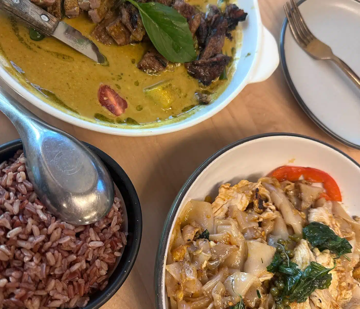 a Top Down View Shows Thai Green Curry with Sliced Grilled Steak a Bowl of Red Rice and a Dish of Sen Thai Noodles with Chicken a Metal Knife Rests in the Curry Bowl and a Fork Sits on a Small Side Plate Atop a Light Wood Table
