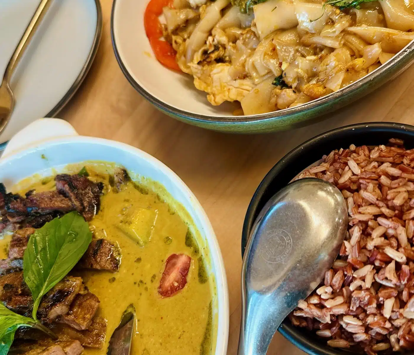 a Spread of Thai Dishes Includes a Bowl of Green Curry with Grilled Meat a Plate of Sen Thai Noodles with Basil and a Side of Red Rice a Metal Spoon Rests in the Bowl of Rice and a Gold Toned Utensil Lies on a Small Empty Plate Nearby