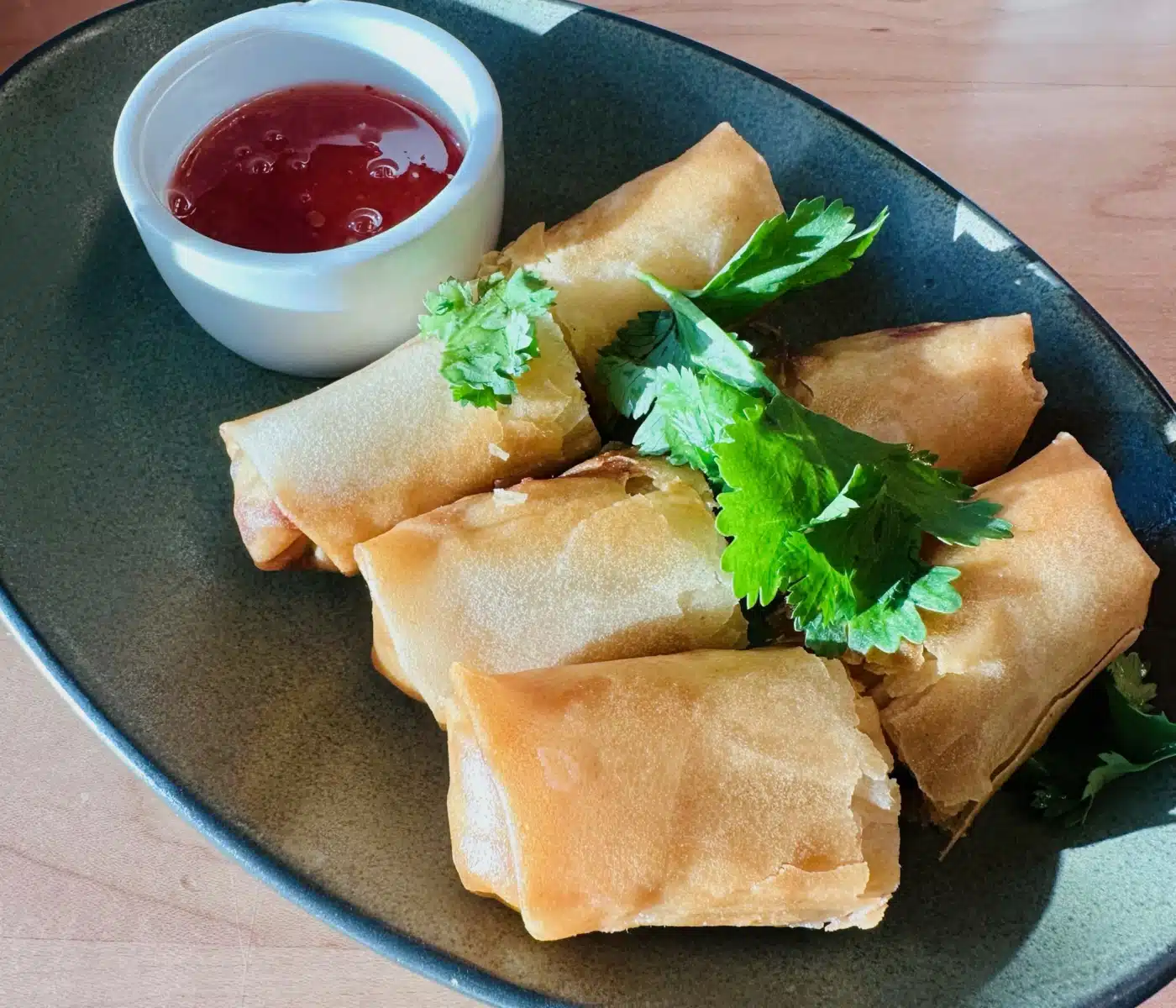 Five Golden Brown Sen Thai Noodles Spring Rolls Are Served on an Oval Plate with a Small Side Dish of Red Dipping Sauce Garnished with Sprigs of Fresh Green Cilantro