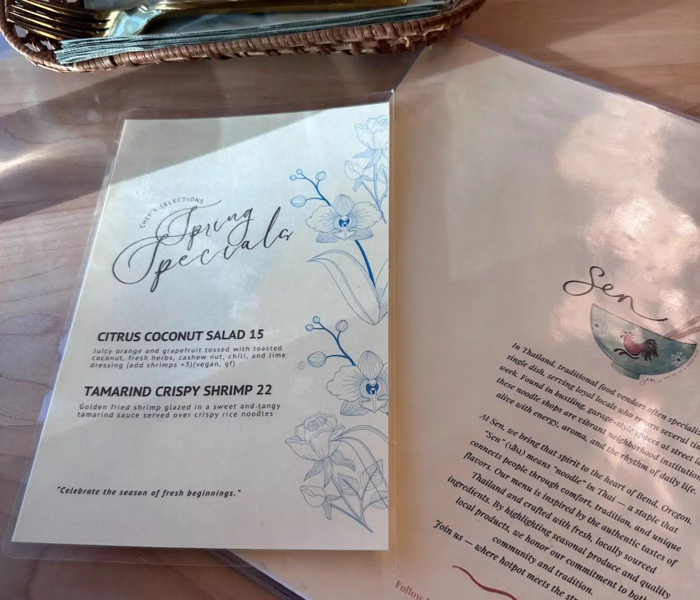 a Sen Thai Noodles Menu Sits on a Light Wooden Table Next to a Woven Basket Containing Gold Cutlery and a Folded Napkin the Menu Features a Spring Specials Section with Two Dishes Decorative Blue Line art Flowers and a Block of Text Describing the Restaurants Background and Mission