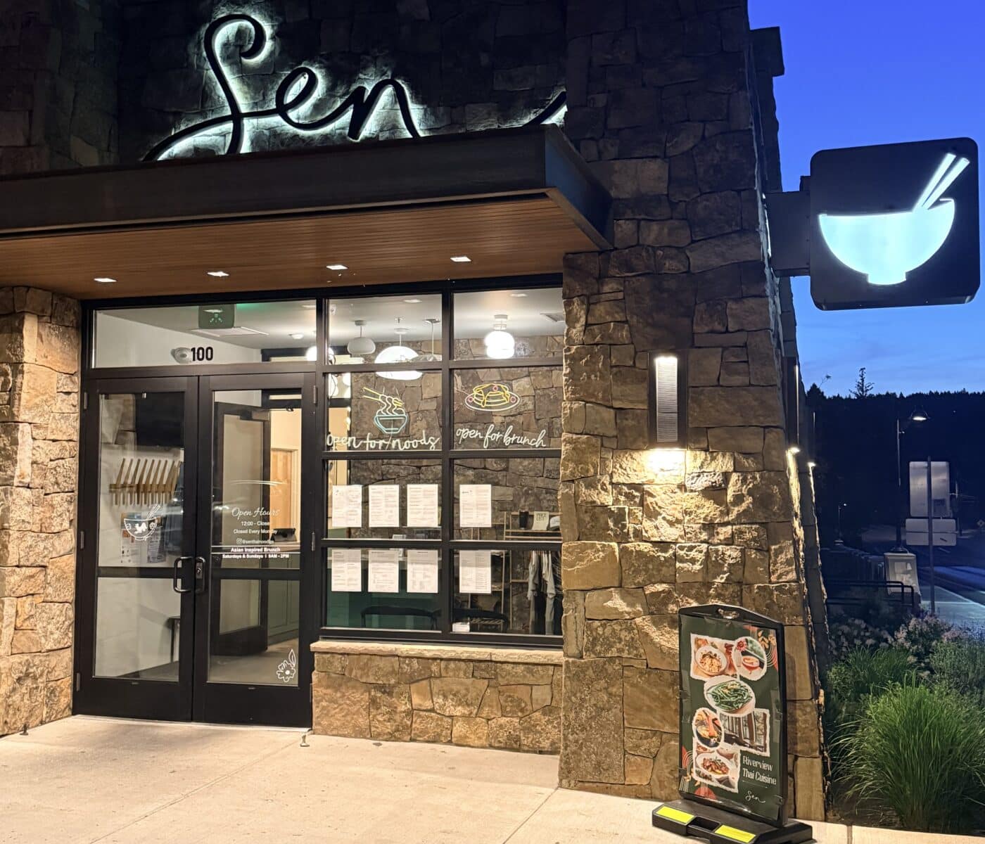 the Stone Faced Exterior of Sen Thai Noodles Features a Glowing Script Sign Glass Double Doors and Window Decals for Noodles and Brunch a Menu Board Stands on the Sidewalk Nearby and a Square Sign with a Bowl Icon Projects from the Corner