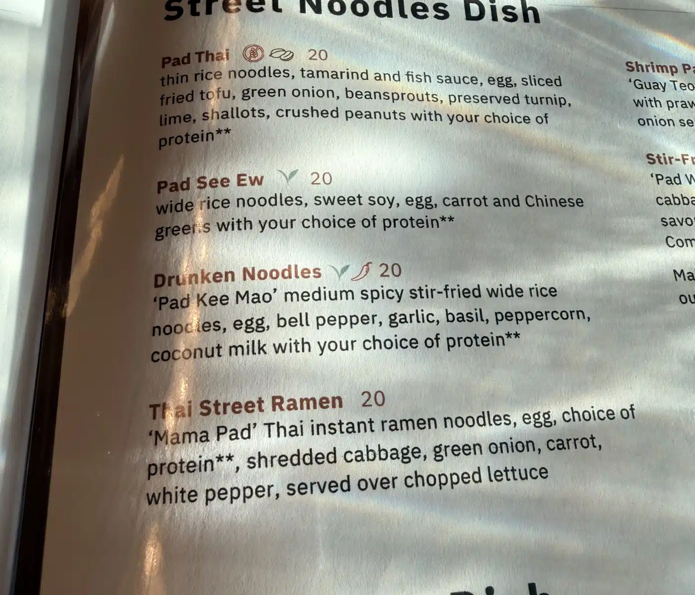 a Close up View of a Sen Thai Noodles Menu Page Lists Street Noodles Options Including Pad Thai Pad See Ew Drunken Noodles and Thai Street Ramen Each Entry Provides a Description of Ingredients and a Price with Some Items Marked by Icons for Vegetarian or Spicy Dishes
