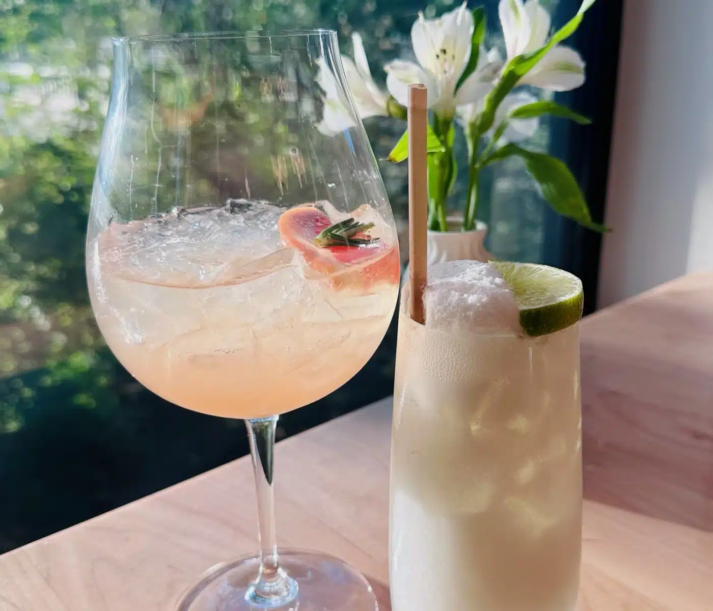 Two Cocktails Sit on a Wooden Table at Sen Thai Noodles in Front of a Window the Glass on the Left Contains a Pink Drink with Ice and a Grapefruit Slice While the Taller Glass on the Right Holds an Opaque White Drink Garnished with a Lime Wheel and a Straw Near a Vase of White Flowers