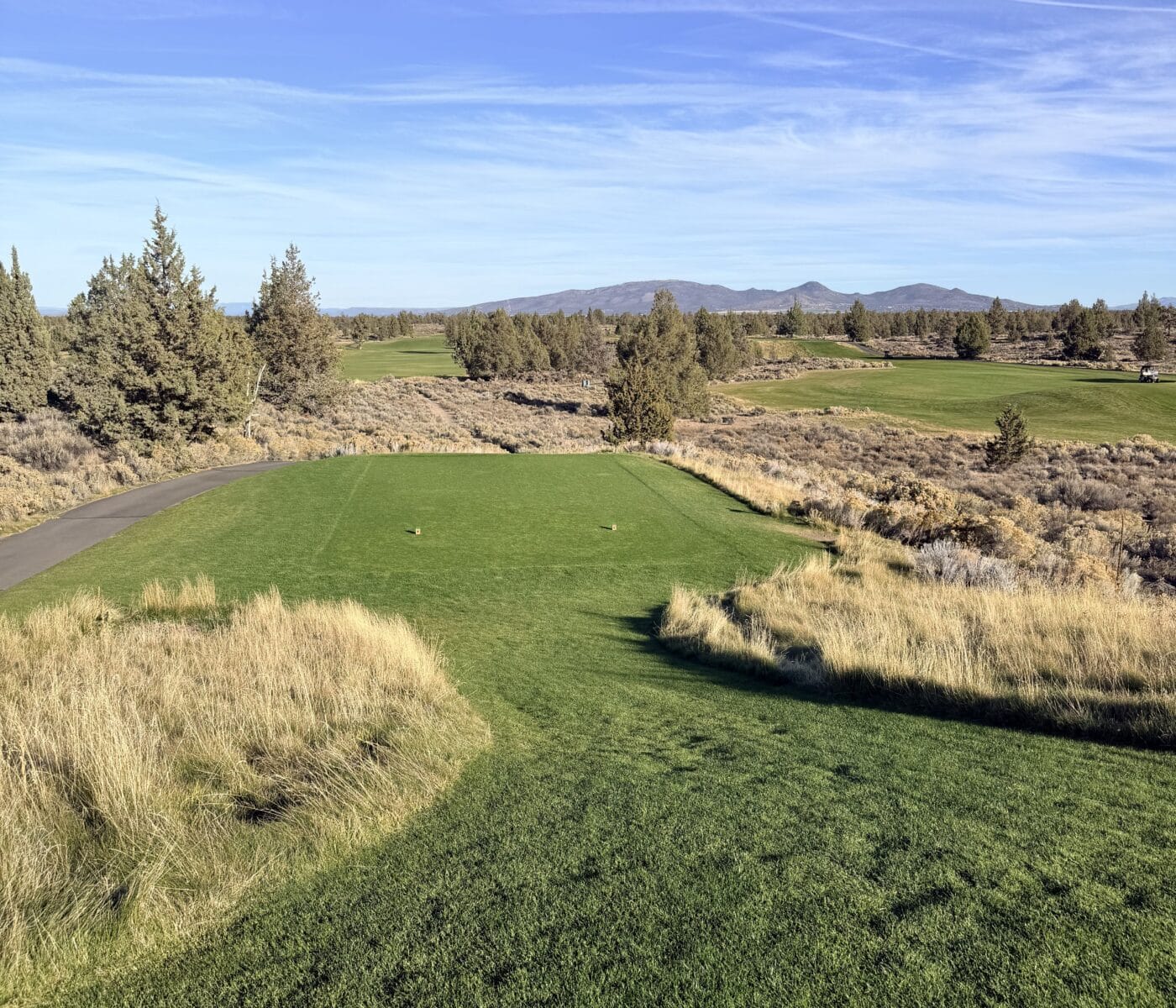 an Elevated View of the Juniper Golf Course Tee Box Overlooks a Rugged Landscape of Dry Golden Grasses and Scattered Evergreen Trees in the Distance Rolling Green Fairways Stretch Toward a Line of Hazy Blue Mountains Under a Clear Bright Sky