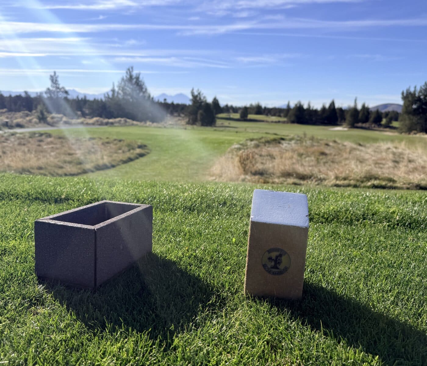 a Rectangular Concrete Trash Bin and a Tall Wooden Tee Marker Sit on a Lush Green Juniper Golf Course in the Background a Sunny Fairway Stretches Toward a Forest Under a Bright Blue Sky with Light Clouds