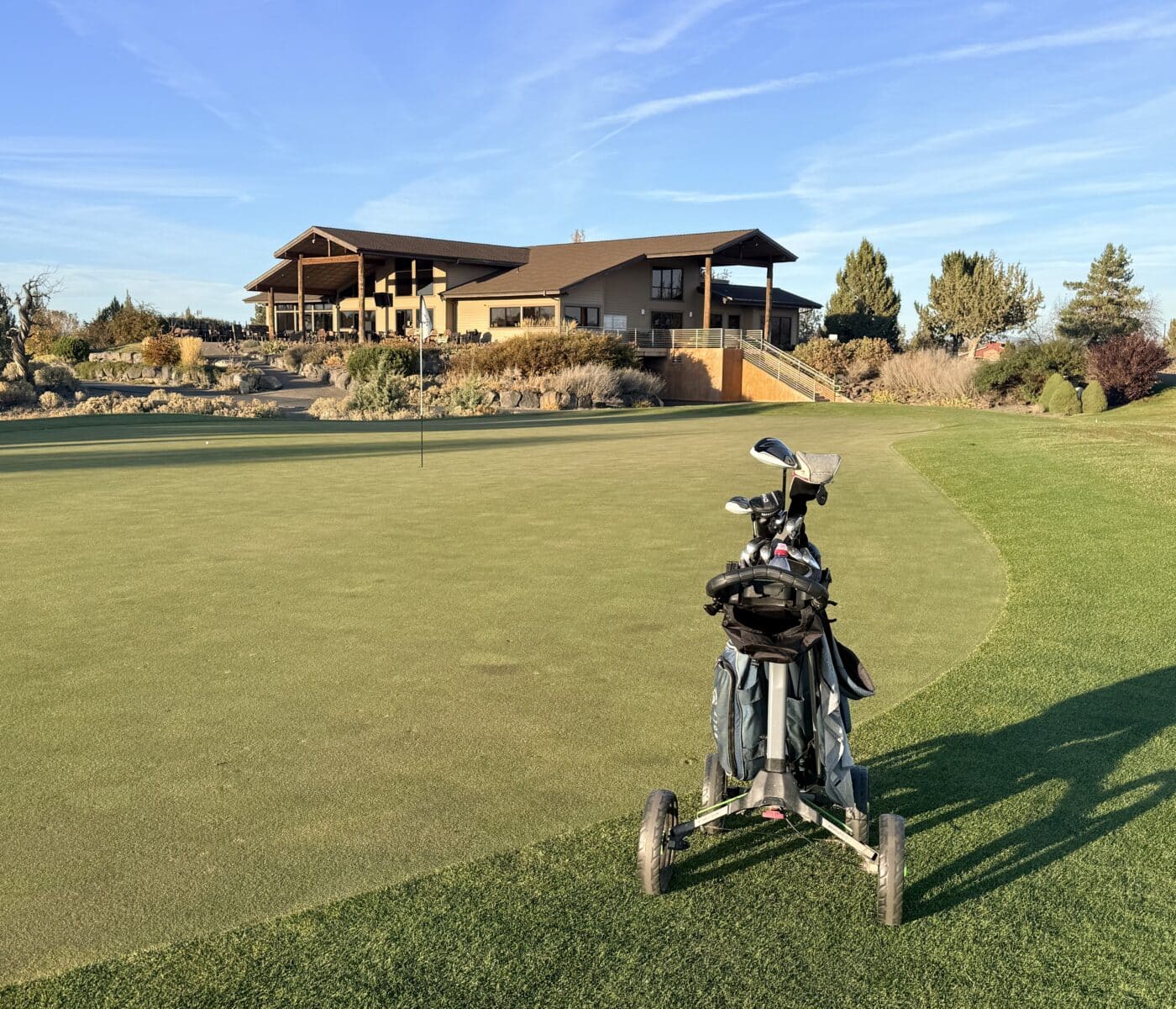 a Golf Bag on a Push Cart Sits on the Edge of a Putting Green at Juniper Golf Course in the Background a Large Modern Clubhouse with Brown Siding and a Patio Sits on a Grassy Hill Under a Clear Blue Sky