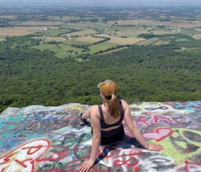 A woman with a ponytail and tan cap sits on a colorful graffiti-covered rock ledge, enjoying one of the best hikes around Frederick MD, as she overlooks green fields and forests under a bright blue sky with scattered clouds.