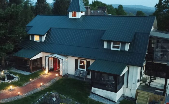 Antietam-Overlook-Farm A large, dark-roofed house with dormer windows and a central peaked tower, resembling the charm of historic hotels in Frederick MD. It features screened porches, garden beds, a red-brick walkway, and green trees under a cloudy sky.