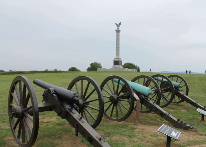 Row of old cannons on a grassy parade ground with a tall monument featuring an eagle statue in the distance