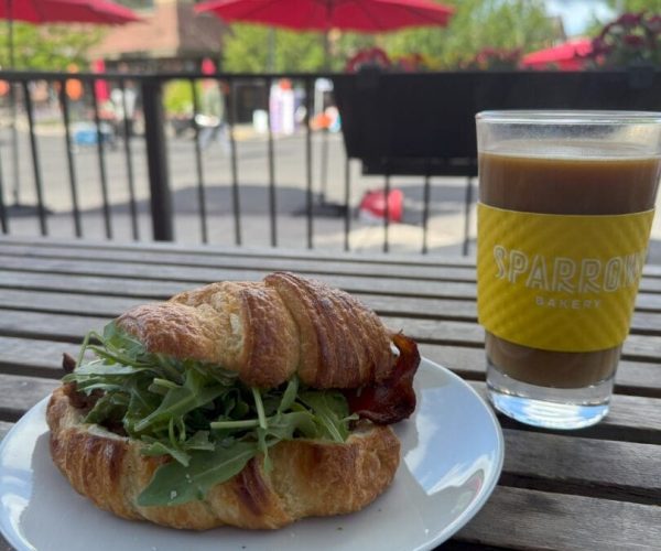 A croissant sandwich filled with arugula sits on a white plate next to a glass of iced coffee labeled Sparrow Bakery, atop a wooden table outside. Red umbrellas, greenery, and buildings are visible in the sunny background.
