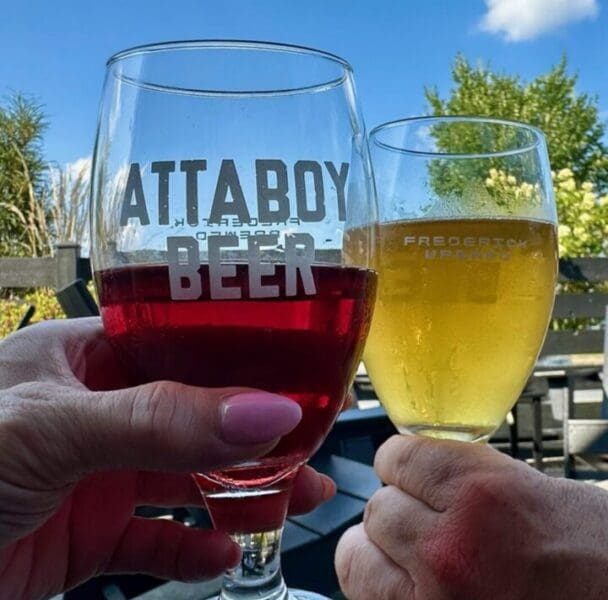 Two hands toast with drinks outside under a blue MD sky—one holding a glass of red beer labeled ATTABOY BEER, the other a yellow beer. A wooden fence, green trees, and patio furniture complete this classic "things to do in Frederick" scene.