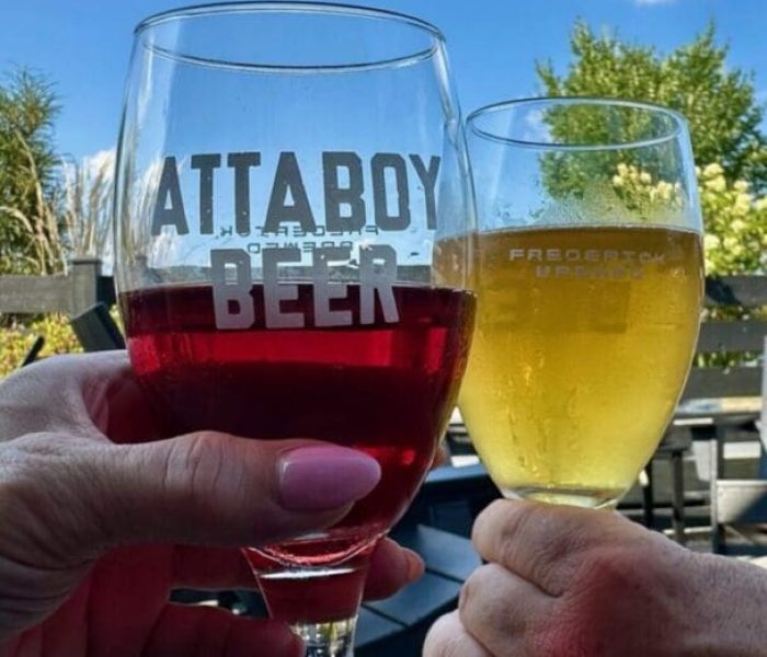 Two hands toast with drinks outside under a blue MD sky—one holding a glass of red beer labeled ATTABOY BEER, the other a yellow beer. A wooden fence, green trees, and patio furniture complete this classic "things to do in Frederick" scene.