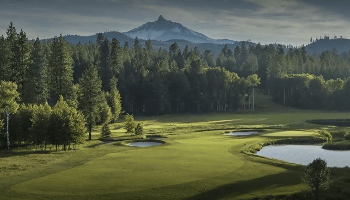 A golf course with green fairway, sand bunkers, and a water hazard, bordered by trees and distant mountains in a serene landscape.