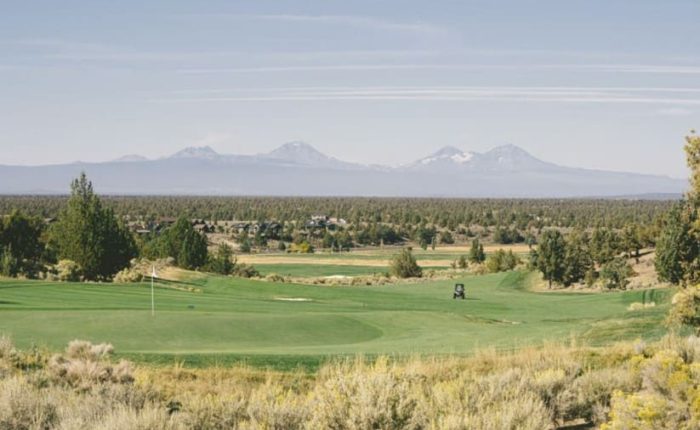 A scenic golf course with lush green fairways and a golf cart sits in the center. Sagebrush and trees surround the course near rolling forests and snow-capped mountains—perfect for those seeking where to stay in Bend, Oregon.