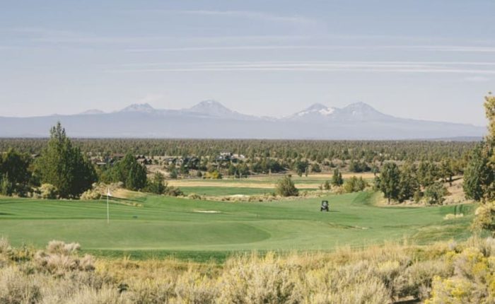 The scenic golf course at Brasada Ranch with lush green fairways and a golf cart sits in the center. Sagebrush and trees surround the course near rolling forests and snow-capped mountains—perfect for those seeking where to stay in Bend, Oregon.