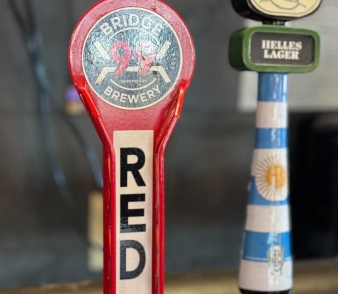 A close-up of two beer tap handles at one of the top breweries in Bend, Oregon. The left handle is red and labeled “RED: BRIDGE BREWERY ROCK CRAWLER.” The right features a cartoon face, green accent, and Argentina flag-inspired stripes.