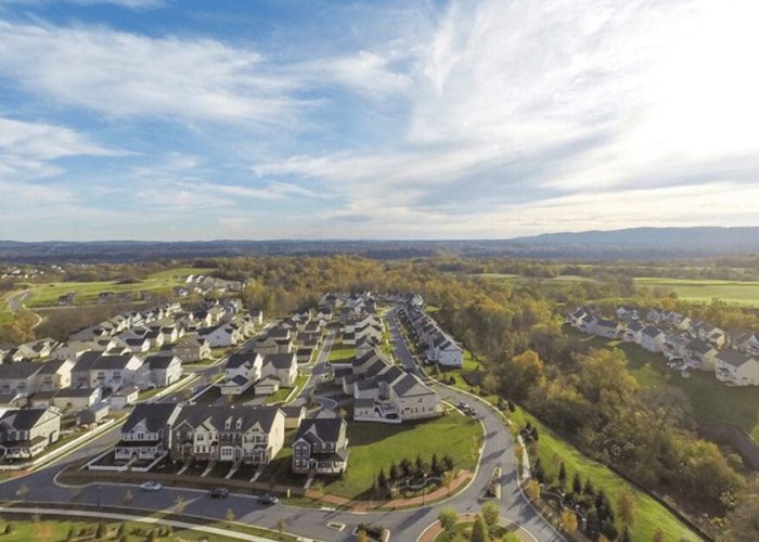 Aerial view of a modern suburban neighborhood with rows of beige houses, curved roads, and green lawns in sunshine.