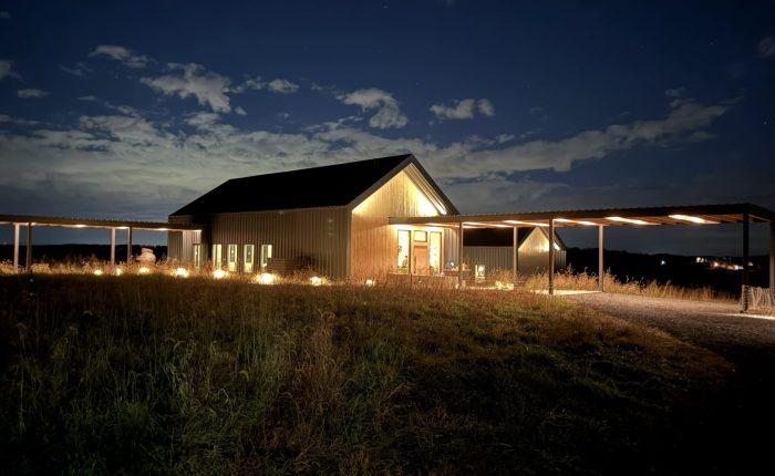 A warmly lit barn-style building stands alone at night under a partly cloudy sky in Mt Airy MD. Soft lights illuminate windows and a covered walkway, while tall grass surrounds the structure, creating a peaceful rural scene.