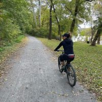A person wearing a black outfit and cap rides a bicycle on a gravel path through a lush, green forest along one of the scenic bike trails near Frederick MD. Fallen leaves border the path, trees line both sides, and a river is partially visible in the background.