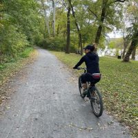 C&O Canal Bike Ride A person wearing a black outfit and cap rides a bicycle on a gravel path through a lush, green forest along one of the scenic bike trails near Frederick MD. Fallen leaves border the path, trees line both sides, and a river is partially visible in the background.