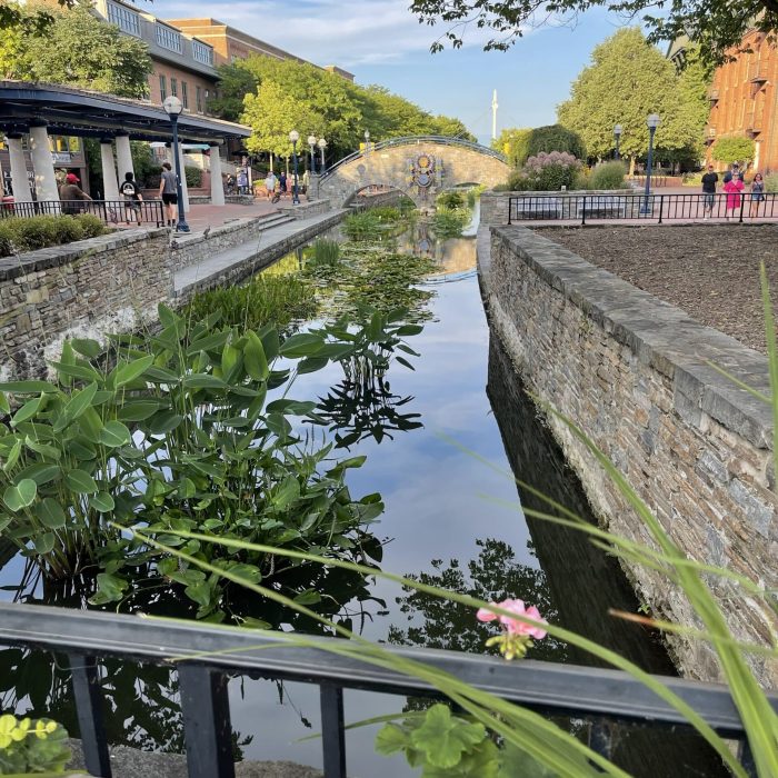 Canal with stone walls and a small arched bridge, pedestrians walking along a tree-lined promenade under a clear sky.