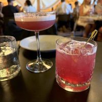 Cocktails-hawkeye-huckleberry Two pink cocktails sit on a dark table: one is in a coupe glass with foam on top, the other in a short glass with ice and a cocktail pick. A glass of water, an empty plate, and people dining inside a warmly-lit tented space are visible in the background.
