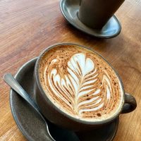 A cup of latte with intricate leaf-shaped art sits on a brown saucer with a spoon, on a wooden table. In the background, an empty matching cup hints at cozy moments found in restaurants in Frederick MD, all warmly lit by natural light.