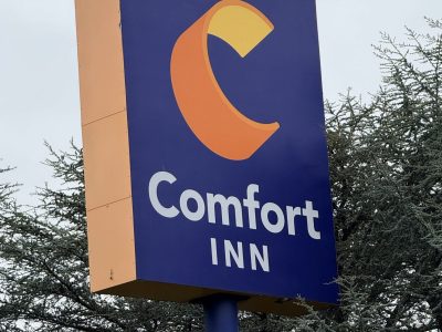 Blue Comfort Inn sign with an orange curved logo on a tall yellow pillar, trees in the background.