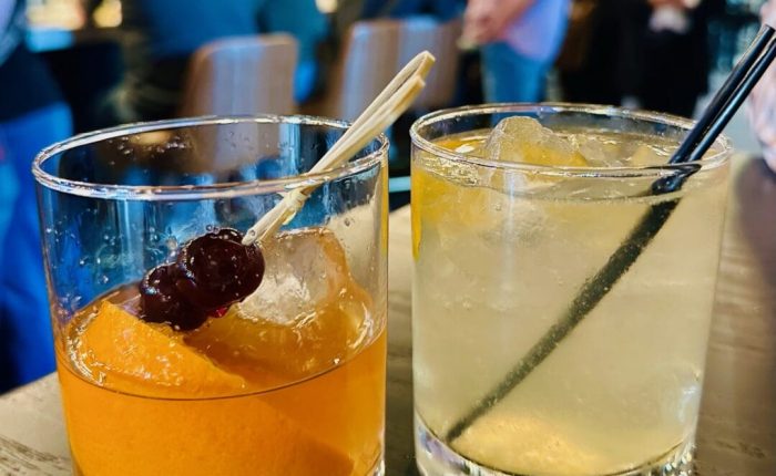Two cocktails in short glasses sit on a black table at a lively spot near a coffee shop in Frederick, MD. One drink features an orange slice and cherries; the other is pale yellow with ice and a black straw. Blurred people and a busy bar fill the background.