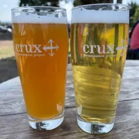Two pint glasses with Crux Fermentation Project logos sit on a wooden table outdoors in Bend, Oregon. One holds a hazy beer, the other a golden brew—perfect for enjoying one of the best things to do in Bend: relaxing with local craft beer.