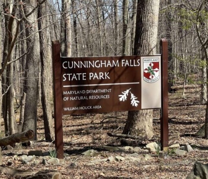 A large wooden sign in a leafless forest marks Cunningham Falls State Park, one of the best hikes around Frederick MD. The Maryland parks logo is on the top right, surrounded by fallen leaves, rocks, and bare trees.