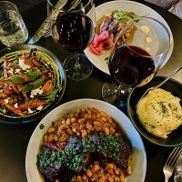 Overhead view of a table set with two glasses of red wine, a plate of stewed beans with braised meat and green sauce, a bowl of creamy mashed potatoes, a dish of roasted carrots topped with mint and feta, and a plate of sliced meat with pickled onions and greens.