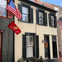 Two-story brick house with dark shutters, white front door, and two American flags plus a red Marine Corps flag outside the window. Wreath on the door.