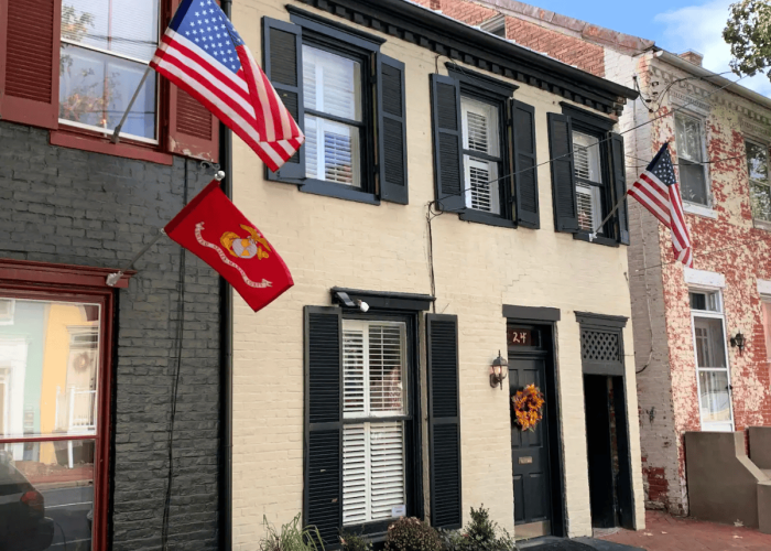 Two-story brick house with dark shutters, white front door, and two American flags plus a red Marine Corps flag outside the window. Wreath on the door.
