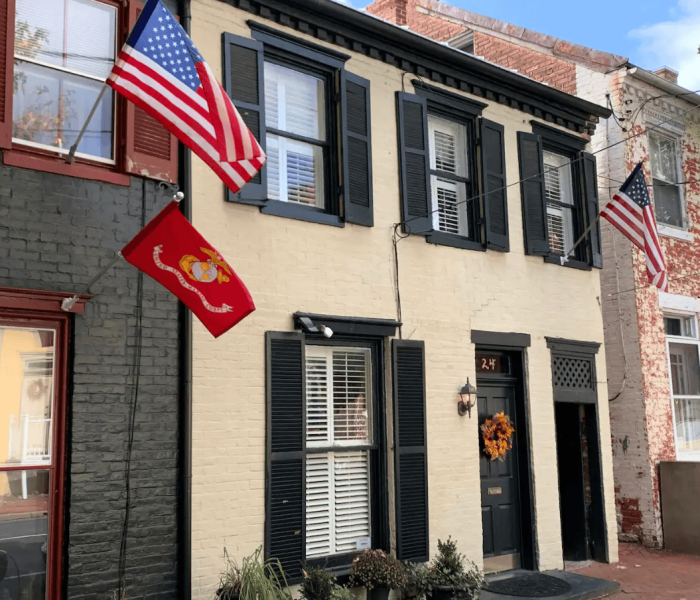 Two-story brick house with dark shutters, white front door, and two American flags plus a red Marine Corps flag outside the window. Wreath on the door.