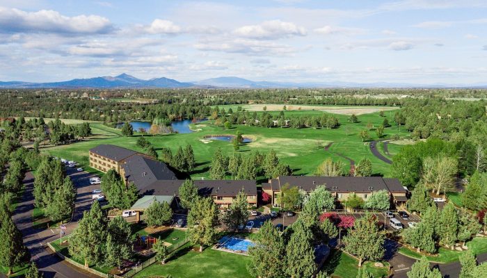 Aerial view of a golf course with green fairways, ponds, and surrounding trees near a hotel building in the foreground.
