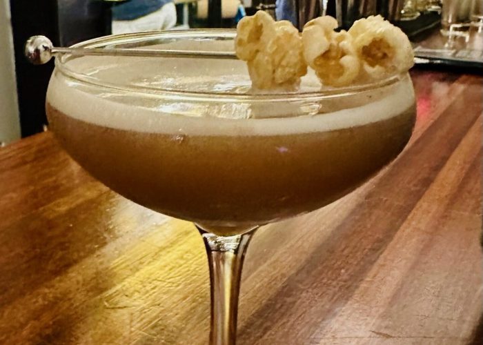 A creamy cocktail garnished with popped popcorn sits in a coupe glass on a wooden bar counter at Cucina Massi Frederick MD. Warm lighting and shelves lined with bottles create a cozy atmosphere as a bartender works in the background.