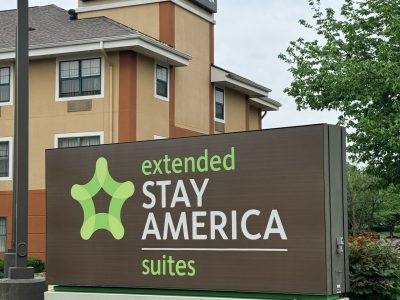 Exterior view of a Stay America sign on a beige building with a green abstract logo and the words 'extended STAY AMERICA'