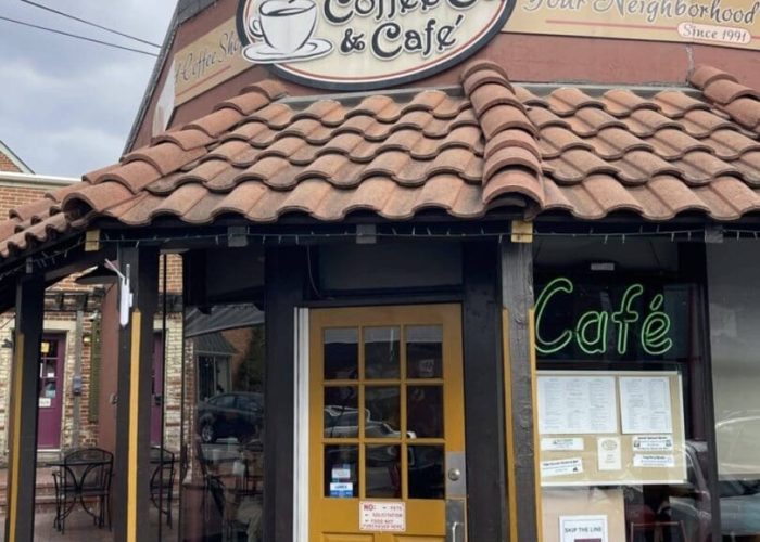 A coffee shop with a brown tile roof, yellow door, large windows, and a sign reading “Frederick Coffee Co & Cafe.” The neon “Cafe” sign glows in a window. Outdoor seating is visible, with tables and chairs along the brick sidewalk—an inviting spot nestled in one of the charming neighborhoods in Frederick, MD.