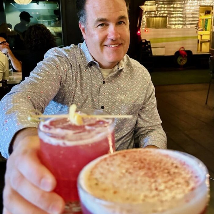 A smiling man in a patterned shirt sits at a table, holding up a pink cocktail with a garnish. Another hand in the foreground holds a similar pink drink, capturing the lively atmosphere of Bend restaurants, with string lights and a shiny food truck behind.