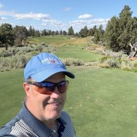 A man wearing a blue cap, sunglasses, and a striped polo shirt smiles for a selfie at Pronghorn Golf Course. Behind him are green fairways, scattered shrubs, pine trees, and a bright blue sky with some clouds.