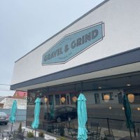 Gravel-and-grind-exterior A modern cafe exterior, part of the vibrant restaurants in Frederick MD, features a sign reading Gravel & Grind above large windows. Closed teal umbrellas shade outdoor tables, with reflections of nearby buildings visible on a cloudy day.