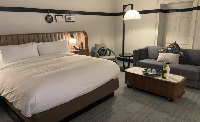 A cozy hotel room in one of the top hotels in Frederick MD, featuring a large bed with a gray headboard, white bedding, a gray armchair, sofa with patterned pillows, wooden coffee table with drinks, wall art above the bed, and a floor lamp.